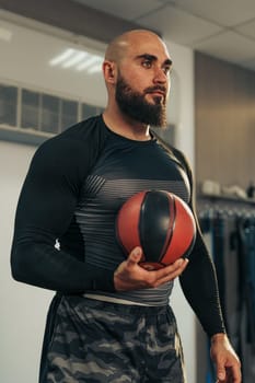 A strong male athlete stands in a gym, holding a basketball while focusing intently on his upcoming training session. He is dressed in a fitted workout outfit, reflecting dedication to sport.