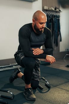 A man with a beard kneels on a gym mat, focused on his workout routine. He wears fitted training gear and demonstrates dedication to fitness in a well-equipped space.