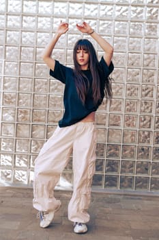 A dancer showcases her skills in an urban environment characterized by a glass brick wall. She wears casual attire and expresses her creativity through movement and style under natural light.