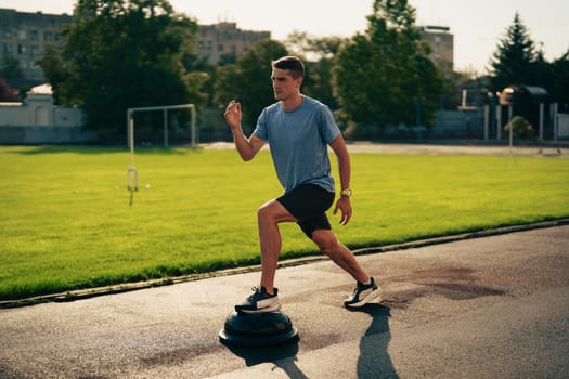 A young man is engaged in balance training on a stability disc at a city park during the late afternoon. The area is surrounded by grass and trees, providing a calm environment.