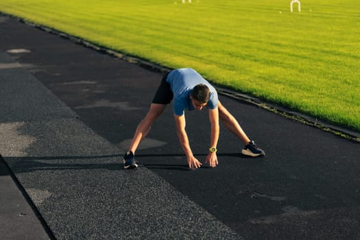 A man performs a stretching exercise on a black running track surrounded by lush green grass during early morning hours. The atmosphere is fresh and energetic.