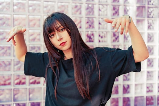 A young woman with long dark hair strikes a playful pose, showcasing her style against a translucent glass wall in a vibrant urban setting during daytime.