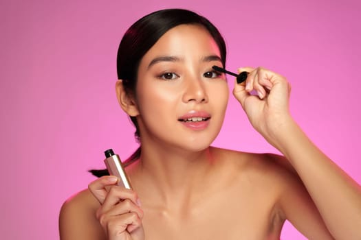 A woman showcases her makeup routine while applying mascara with precision. She smiles confidently, illuminated by soft lighting in a beauty studio, enhancing her natural features.