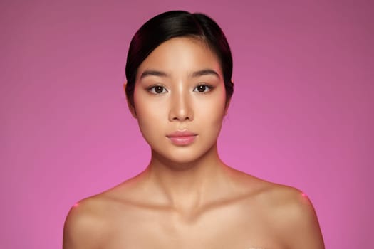 A woman stands confidently with a serene expression, showcasing natural beauty. The vibrant pink background adds a contemporary touch, enhancing her features.