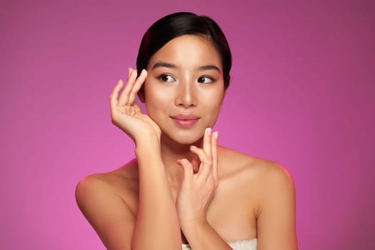 A model gracefully poses with her hands near her face, displaying a serene expression. The vibrant pink backdrop emphasizes her features in a professional studio setting.