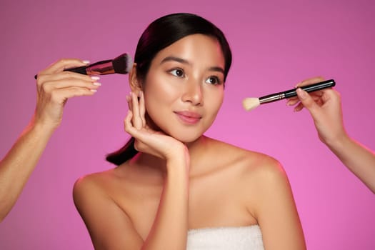 Two hands apply makeup to a model with smooth skin, highlighting beauty techniques against a vivid pink background. The atmosphere feels professional and vibrant.