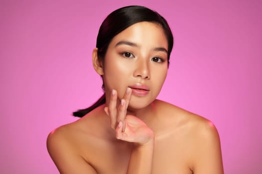A model showcases her serene expression and flawless skin while gently touching her face against a vibrant pink background. The lighting emphasizes her features.
