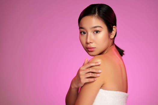 A model poses gracefully with a soft expression, lightly touching her shoulder while surrounded by vibrant pink lighting in a studio setting, highlighting her natural beauty.