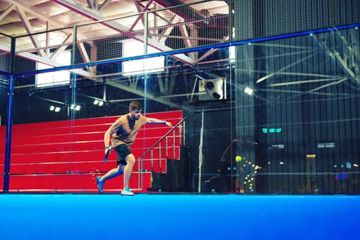 A focused male athlete is engaged in a fast-paced squash match inside a contemporary sports center. The blue court and vibrant red seating create a dynamic atmosphere.