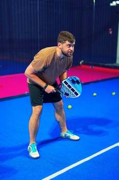 A man stands in an indoor padel court, focused and ready to play. He holds a padel racket with a determined expression, surrounded by yellow balls on a vibrant blue surface.