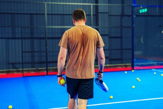 A young man walks towards the court, holding a padel racket and ball after practice. Sweat on his shirt highlights his dedication to the sport in an indoor setting.