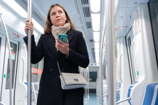 Young woman in a black coat and beige scarf standing inside a modern subway train, holding a smartphone and gripping a metal pole. She looks thoughtful while commuting in the city.