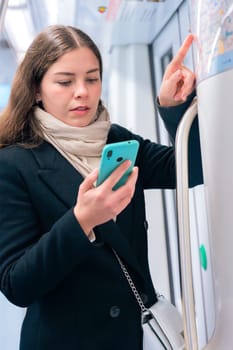 Focused young woman in a black coat and beige scarf checking a subway map while holding her smartphone. She navigates her route inside a modern metro train.