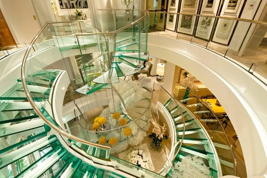 Modern spiral glass staircase in luxury interior with artwork and chandeliers.