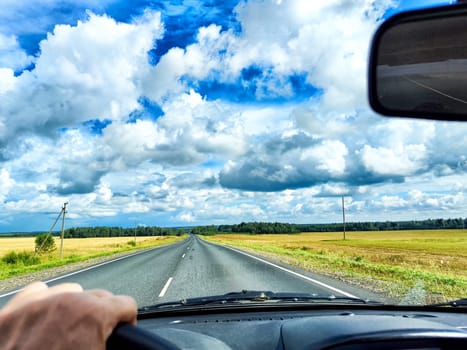A hand grips the steering wheel while driving along wide road surrounded by fields under a beautiful blue sky