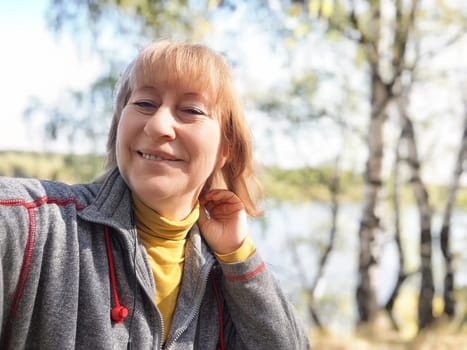 Blonde woman takes a cheerful selfie on a tranquil nature trip in autumn by a picturesque lake and vibrant fall foliage.
