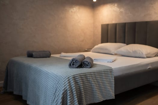 Neatly made double bed with gray headboard, light blue blanket, and rolled towels. Textured wall and dim overhead lighting.