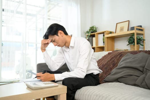 Young man sitting on a couch looking worried while calculating expenses in living room.