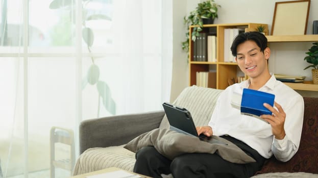 Smiling asian man holding a savings passbook and checking his financial transactions on digital tablet.