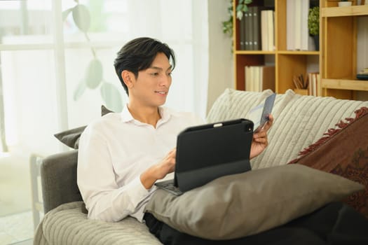 Smiling asian man holding a savings passbook and checking his financial transactions on digital tablet.