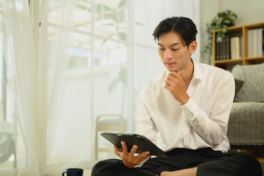 Young asian man browsing or working on digital tablet in living room.