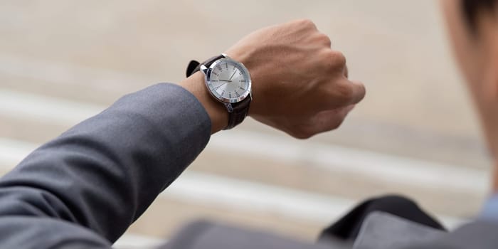 A close up view of a young Asian businessman in a suit, checking his watch, reflecting professionalism and time management in a contemporary outdoor setting.