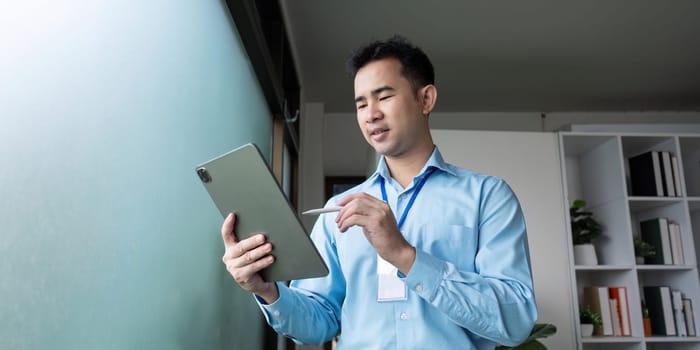 A focused Asian businessman is using a tablet for planning, standing by a window in a contemporary office setting.