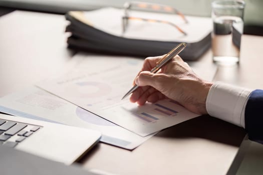 An elderly businessman focused on analyzing financial reports and charts, jotting down notes at his desk in a contemporary office.