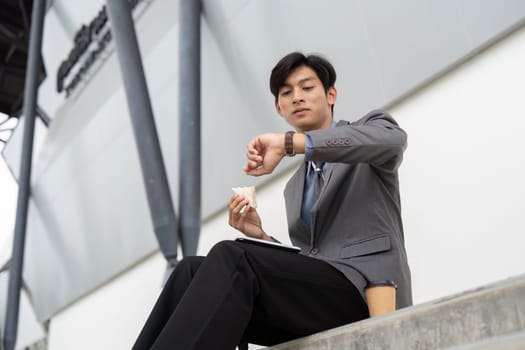 A young Asian businessman in a suit, checking his watch while eating a snack, balancing work and leisure in a contemporary outdoor environment.