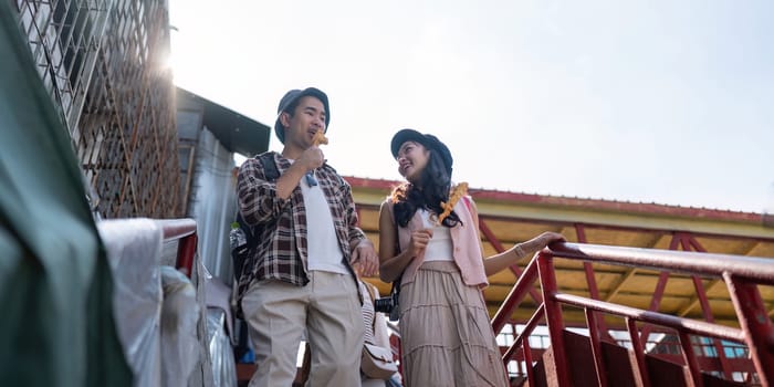 Two happy Asian friends savor street food while walking down a vibrant bridge, sharing smiles and memorable travel experiences.