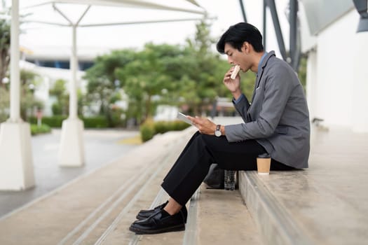A young Asian business professional sits outdoors, eating a snack and using a tablet, showcasing a blend of work and relaxation in a modern environment.