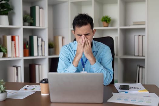 An anxious Asian businessman shows signs of stress at his desk, overwhelmed by work while looking at his laptop and scattered documents in a contemporary workspace.