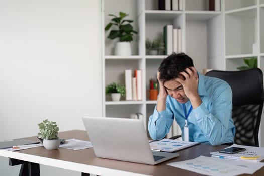 A frustrated Asian businessman holds his head in despair at his desk, struggling with work challenges while surrounded by a laptop and documents in a contemporary workspace.