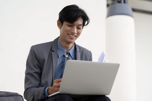 A young Asian businessman in a suit, smiling as he works on his laptop, showcasing a positive and productive atmosphere in a contemporary outdoor setting.