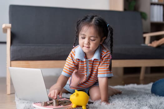 A young girl happily interacts with a laptop while exploring saving money concepts, surrounded by coins and a piggy bank in her warm living space.
