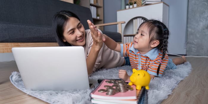 A diverse mother and her daughter celebrate with a high five as they engage in saving money using a yellow piggy bank and laptop, promoting financial literacy at home.