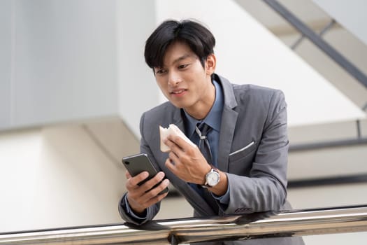 A young Asian businessman casually eating a snack and using his phone in a contemporary office environment, balancing work and personal time.