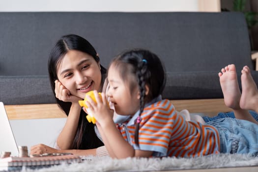 A diverse mother watches her daughter joyfully interact with a yellow piggy bank while they explore financial concepts together using a laptop at home.