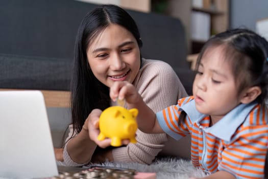 A mother beams with pride as her daughter adds coins to a piggy bank, both engaged in a delightful learning experience about saving money.