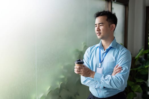 A contemplative Asian businessman stands with arms crossed, holding a coffee cup by a frosted glass window in a contemporary office environment.