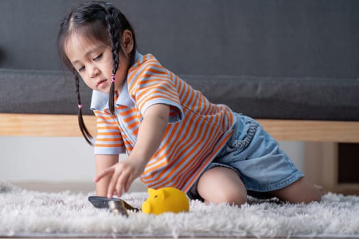 A young girl explores saving money by reaching for a yellow piggy bank and coins on the floor, promoting financial skills in a family setting.
