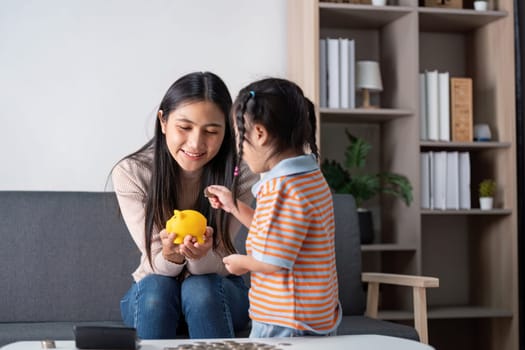 A diverse mother and her daughter share moment of joy as they explore saving money together using yellow piggy bank in their living room.