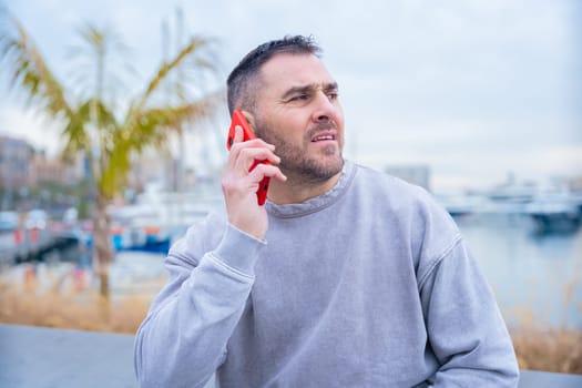 Casual man in a gray sweatshirt talking on a red smartphone near a marina. He appears focused while looking into the distance. Boats and waterfront setting in the background. Modern lifestyle