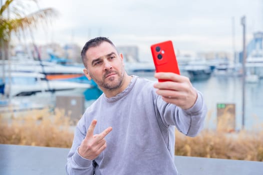 Casual man in a gray sweatshirt taking a selfie with a red smartphone near a marina. He makes a peace sign while posing. Boats and waterfront scenery in the background. Modern lifestyle