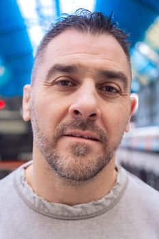 Middle-aged man wearing a gray sweatshirt standing at a train station with a thoughtful expression. The urban background and blue ceiling create a modern and reflective atmosphere.