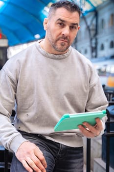 Middle-aged man in a gray sweatshirt sitting at a train station, using a turquoise tablet. His serious expression and urban backdrop suggest work, travel, or digital connectivity.