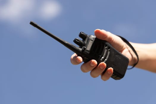 A portable walkie-talkie against a blue sky during a mountain adventure, held in a hand. Emphasizes communication, safety, and readiness in the wilderness. For tourism, extreme sports, teamwork, and outdoor technology themes. Dynamic composition with sunlight and fresh air.