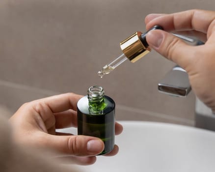 Female hands with opened green dropper bottle and pipette in fingers, Close up. Woman in bathroom wearing brown bathrobe with serum or cosmetic essence