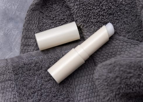Opened white blank lip balm stick on dark grey folded towels top view, cosmetic packaging mockup, copy space. Lipcare beauty product for mens everyday personal care
