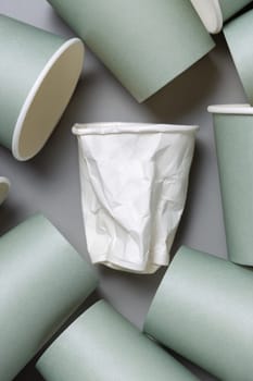 Disposable new light green paper cups and crumpled used white one on gray top view, mockup with copy space. Empty cardboard coffee cups flat lay. Consumerism concept